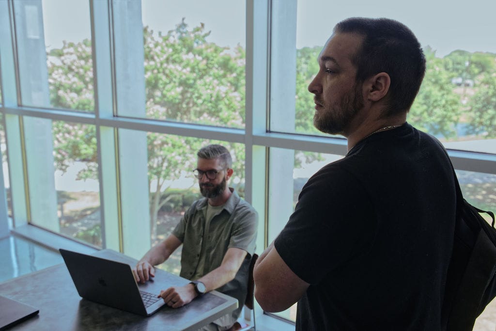 Two professionals working in a modern office with large windows, collaborating on a laptop.