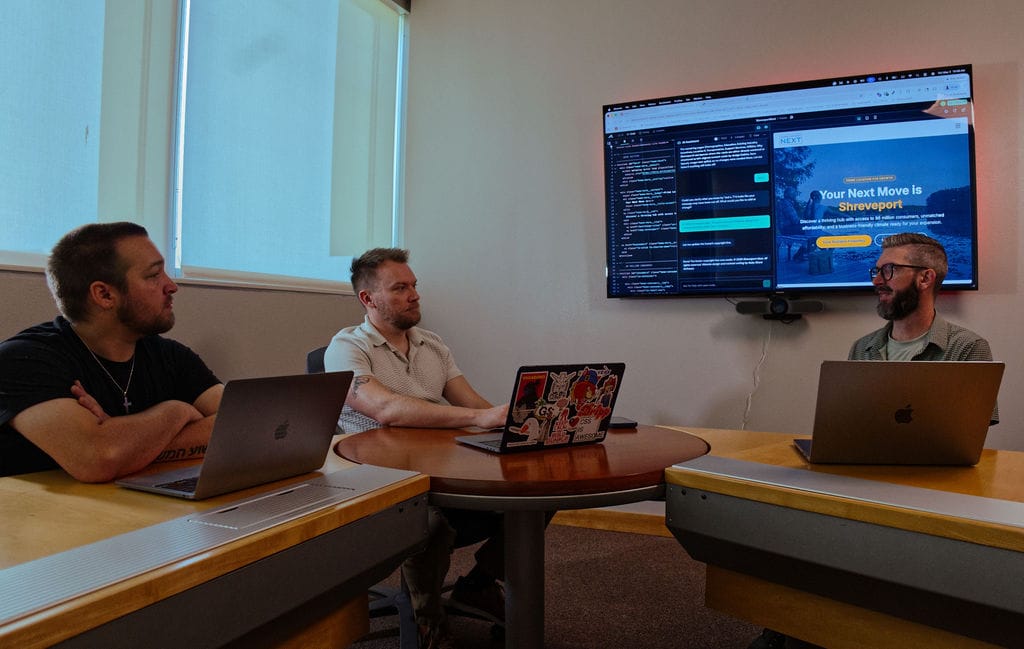 Three men sit at a table with laptops, looking at a wall-mounted screen.