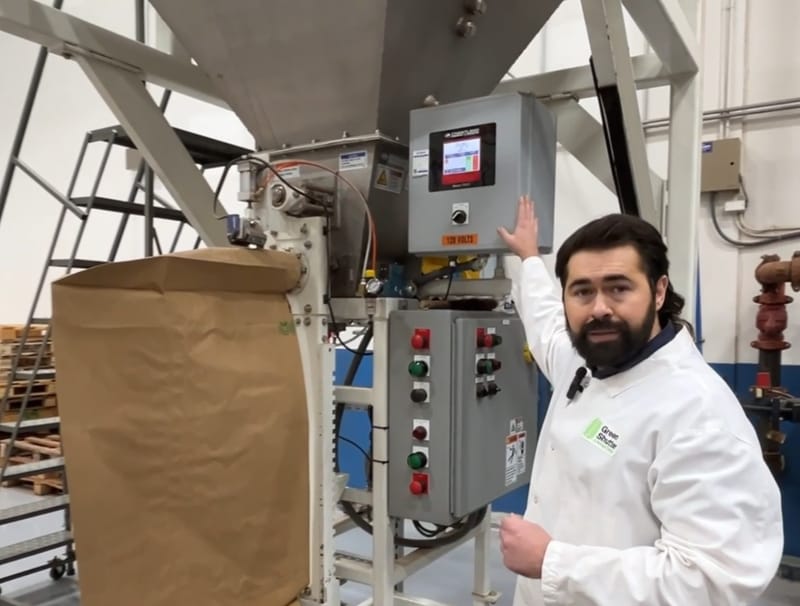 A man in a white coat explaining industrial packaging machinery with controls and a large paper bag.