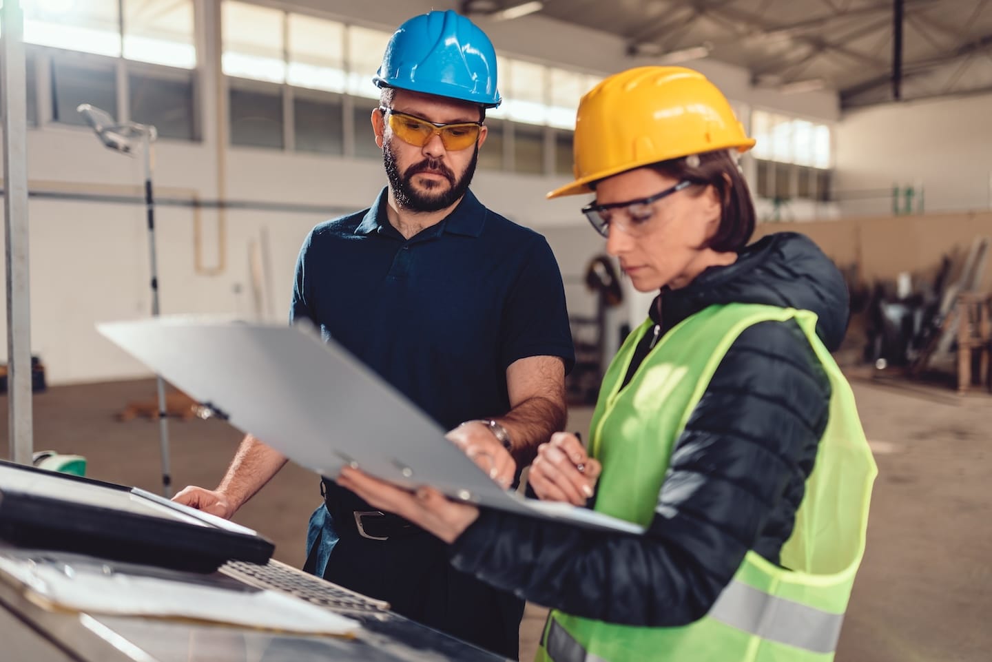 Two construction workers in hard hats reviewing documents on a clipboard in a warehouse