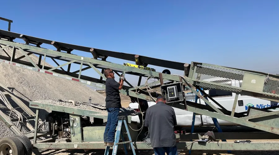 Two workers repairing an industrial conveyor belt system outdoors at a quarry facility