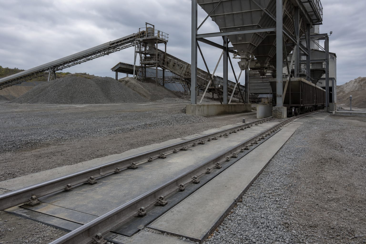 Heavy-capacity rail scale at an aggregate quarry site with hopper bins and conveyor structures in the background
