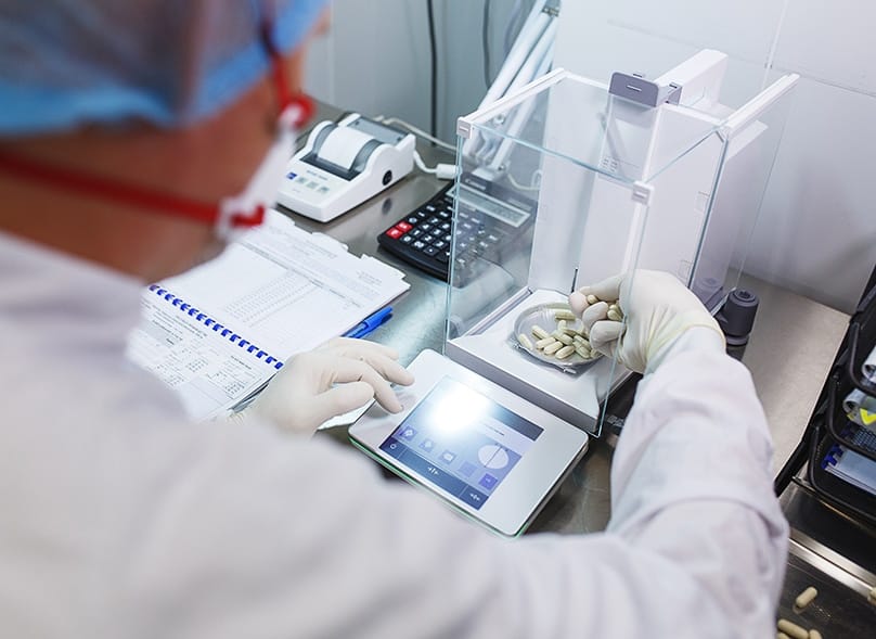Technician in lab attire weighing capsules on a digital scale.