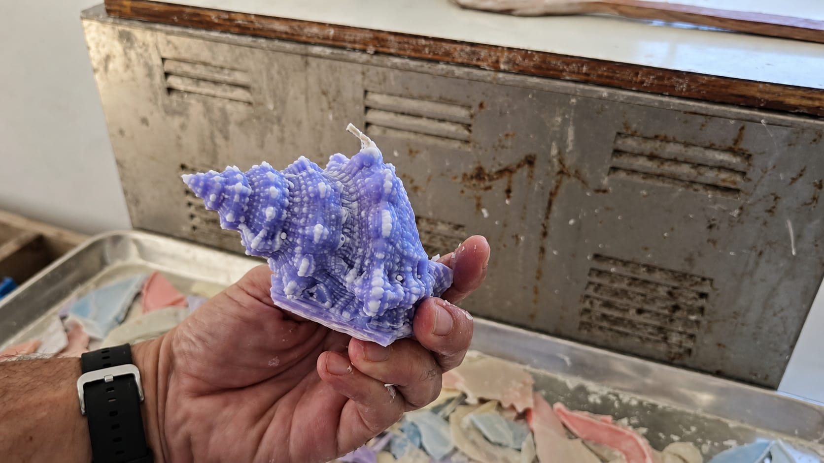 A maker holds a purple-and-white sculptural shell candle during the finishing and inspection process.