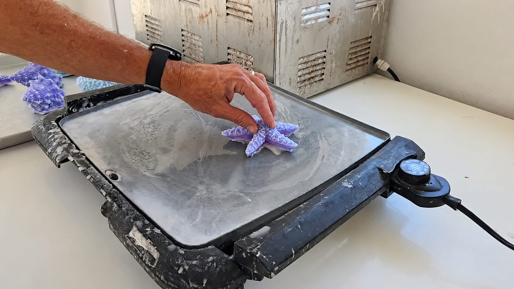 A maker hand-finishes a purple-and-white starfish candle on a heated flat work surface.