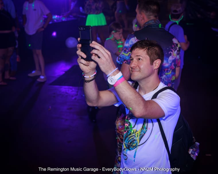 A man in a white T-shirt takes a selfie at a neon-themed party