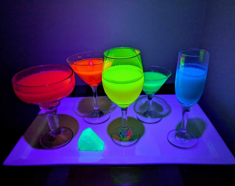 Five glasses with neon-colored drinks glowing under a blacklight